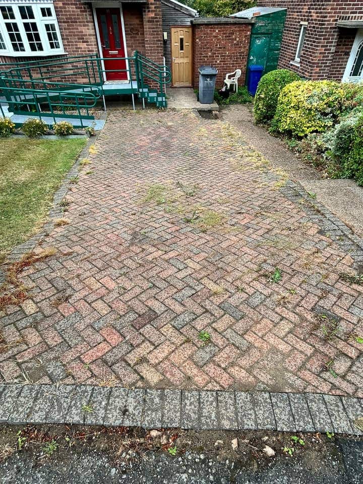 Dull block-paved driveway with weeds along the joints, leading to a house with a red front door