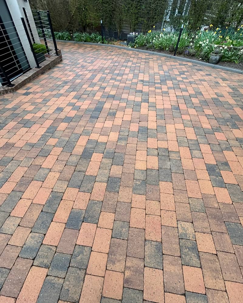 A wide brindle block-paved drive after cleaning, with a flower border and hedge behind