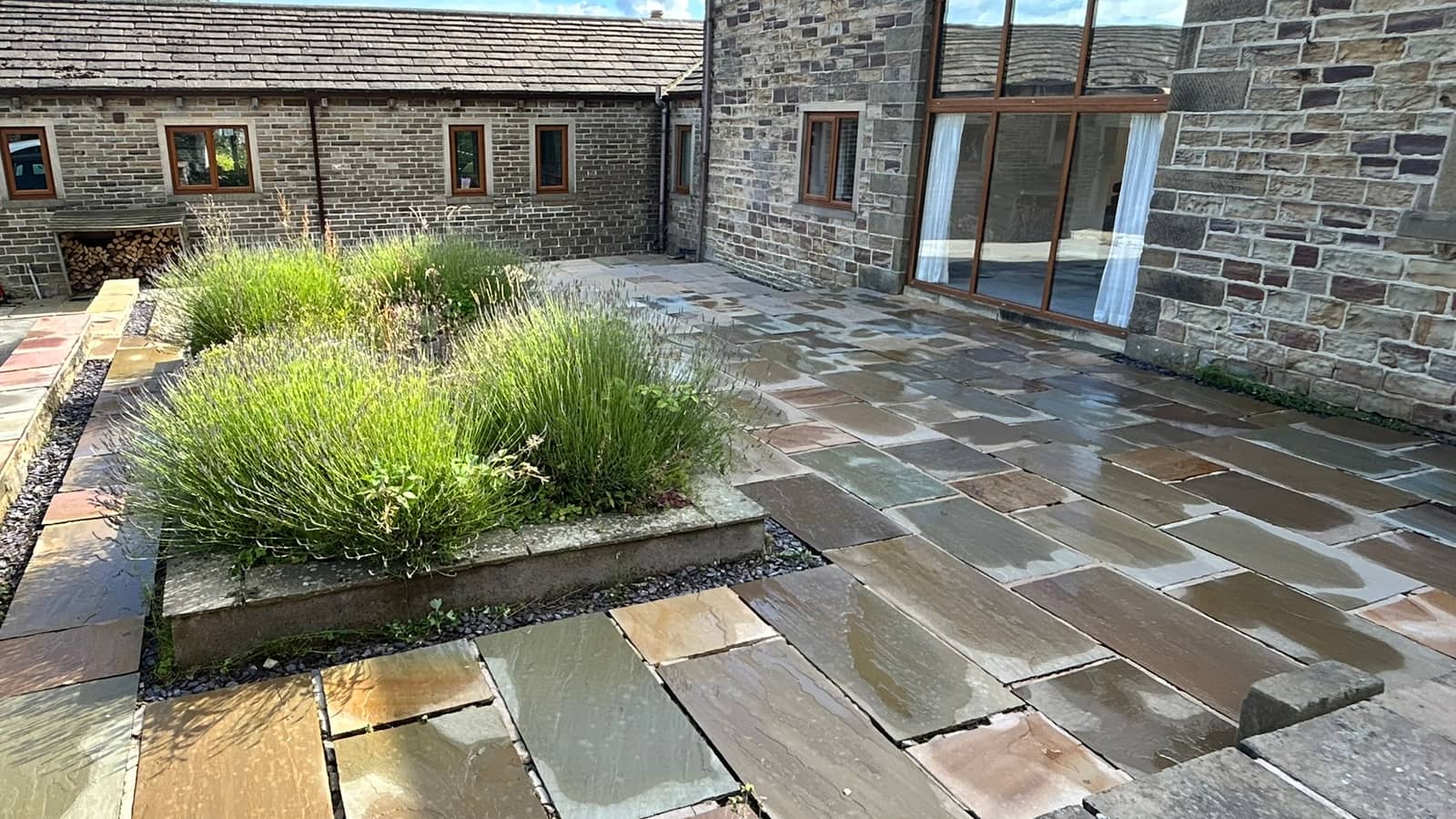 A wet, freshly cleaned Indian stone courtyard with a raised planted bed, in front of a stone barn conversion