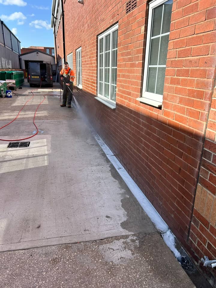 An operative in orange hi-vis washing the base of a red brick wall, the cleaned strip of concrete bright beside it