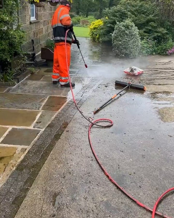 A stone path being pressure washed: an operative in orange hi-vis with a lance, spray rising off the wet stone, a red hose running across the ground
