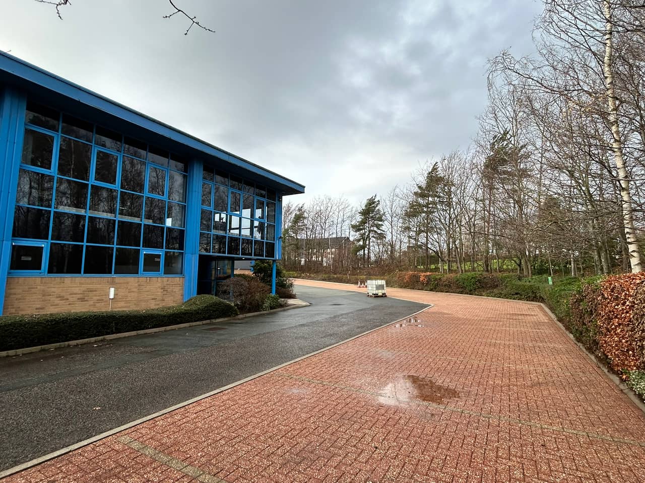 A cleaned red block-paved car park beside a blue glazed office building, the access road still wet