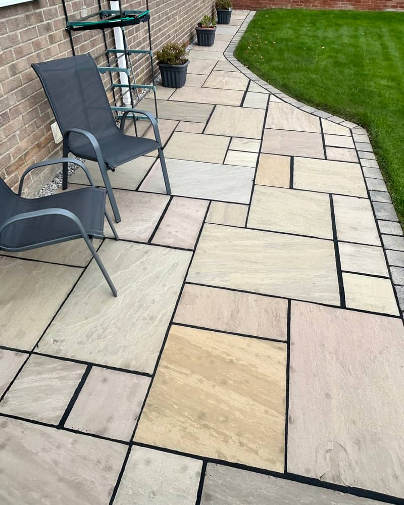 A clean buff Indian stone patio with dark pointing, two garden chairs and a curved lawn edge