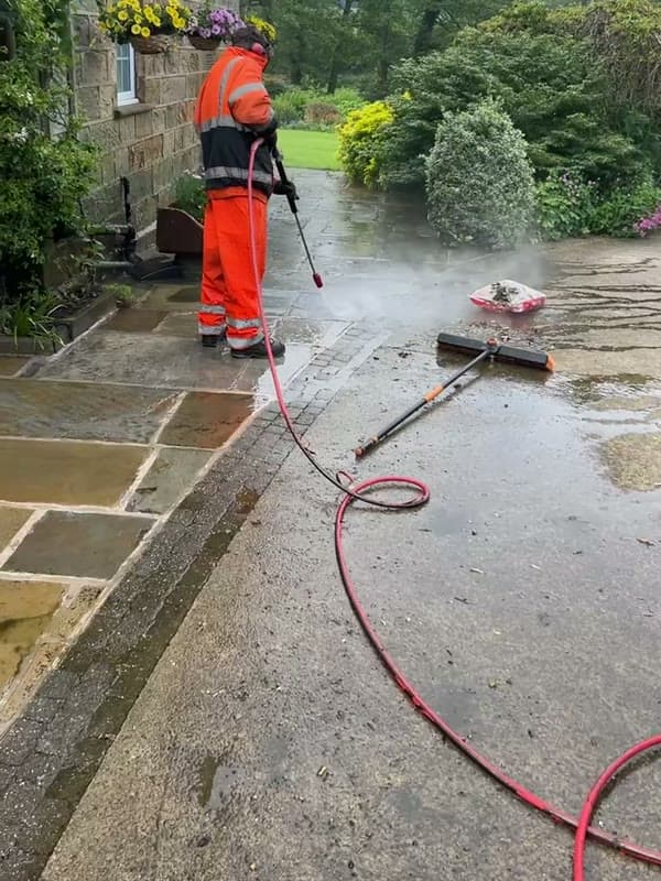 An operative in orange hi-vis pressure washing a stone path, spray lifting off the wet surface
